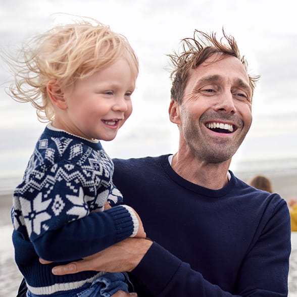 Man walking across a windy beach while carrying a young child in his arms.