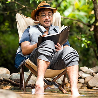 Man relaxing outdoors by a stream while sitting in a foldable chair with his socks off, trousers rolled up to his knees and his feet in the water.