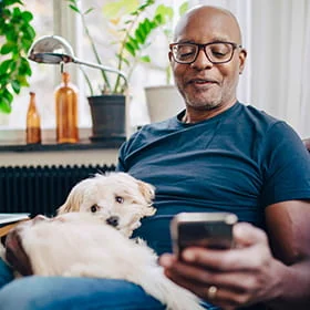 Man sitting relaxed in his living room with his dog on his lap and scrolling through his mobile phone.