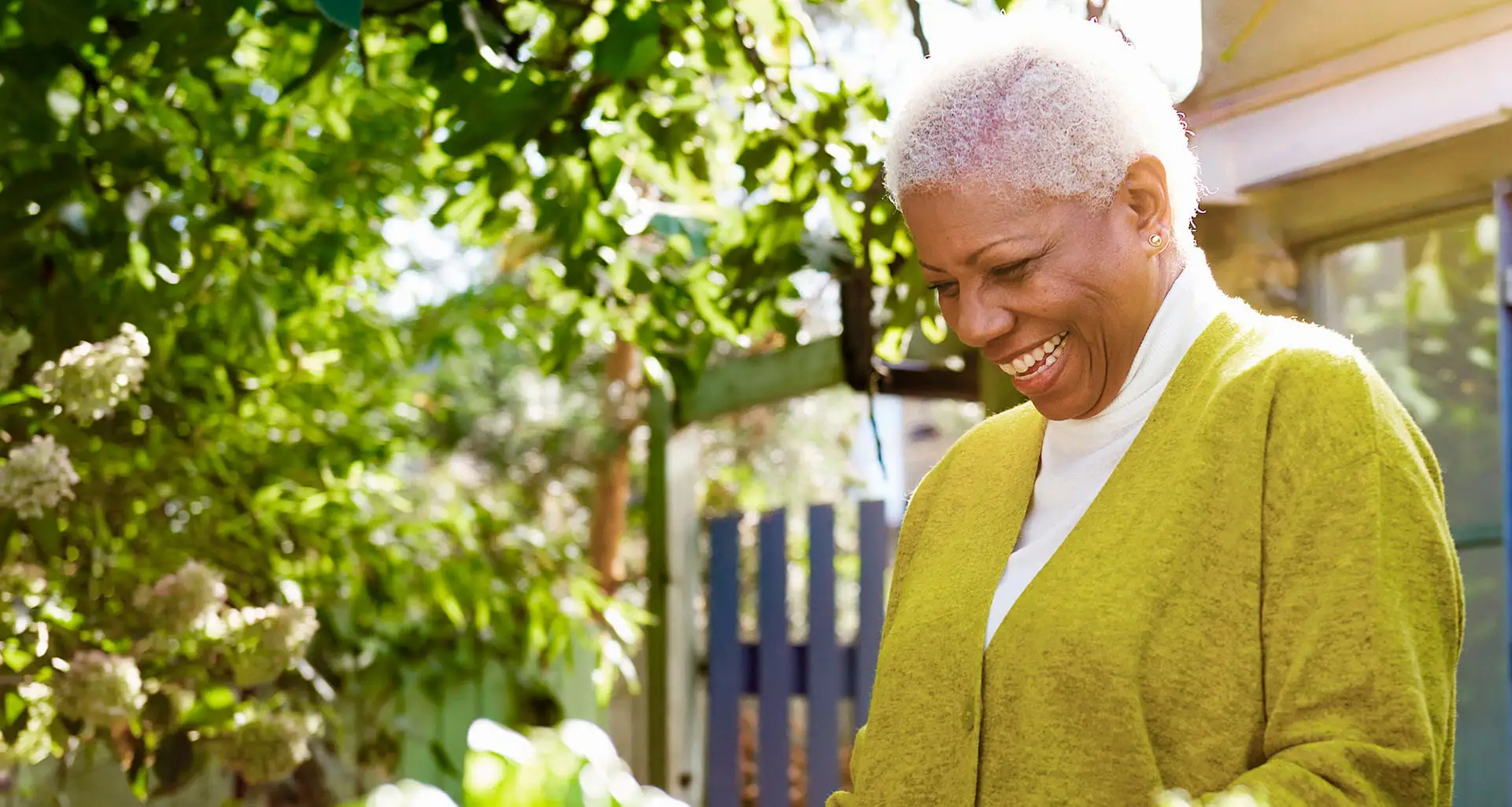 Woman standing in the sunshine outside her home and smiling while tending to her green garden.